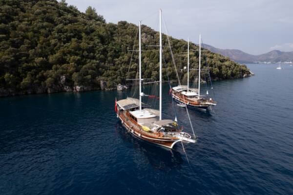 Two beautiful gulet yachts, one identified as Gulet Yucebey 1, cruising side-by-side on the sparkling Turkish waters with lush green mountains in the background.